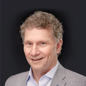 Craig Compiano, a middle-aged man with curly gray hair, smiles confidently in a suit and blue shirt before a plain black backdrop.