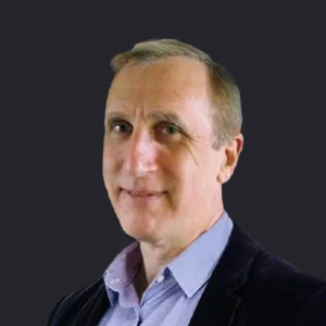 Craig Compiano, middle-aged with short light brown hair, smiles in a blue shirt and dark blazer against a plain dark background.