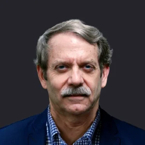Craig Compiano, mature male with gray hair and mustache, in business attire, stands confidently before a solid dark background.